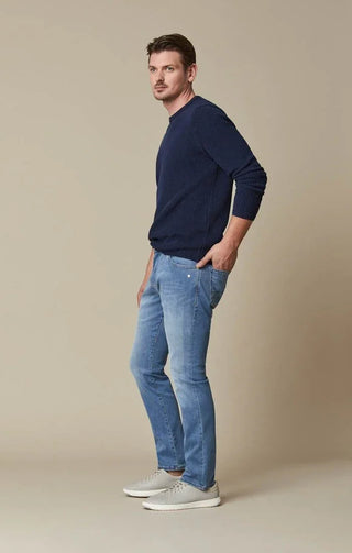 Man wearing light blue slim-fit jeans, navy sweater, and grey sneakers, standing sideways