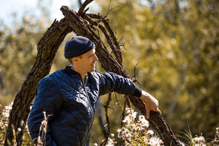 Man outdoors wearing navy waterproof wool beanie and quilted jacket, leaning on a tree