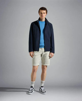 Man wearing navy nylon jacket, blue sweater, beige shorts, and sneakers against plain background
