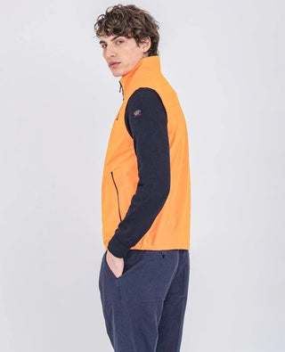 Man wearing sleeveless orange nylon vest, navy sweater, and blue pants, side view